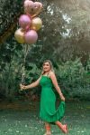 Joyful woman in green dress celebrating her 30th birthday with balloons in a lush outdoor setting.
